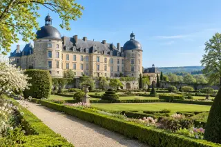 Château de Hautefort sous le soleil de printemps vu depuis ses jardins à la française, façades en pierre blonde, topiaires symétriques, parterres fleuris et pelouses bordées d’arbres sur fond de campagne vallonnée.
